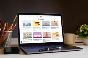 Foto van een opengeklapte laptop. Op het scherm zie je de leidraad toegankelijk bouwen: een aantal bouwstenen in de vorm van gekleurde blokken met illustraties en titels. Naast de laptop een houder met potloden, een telefoon, een koffiebeker en een plantje.